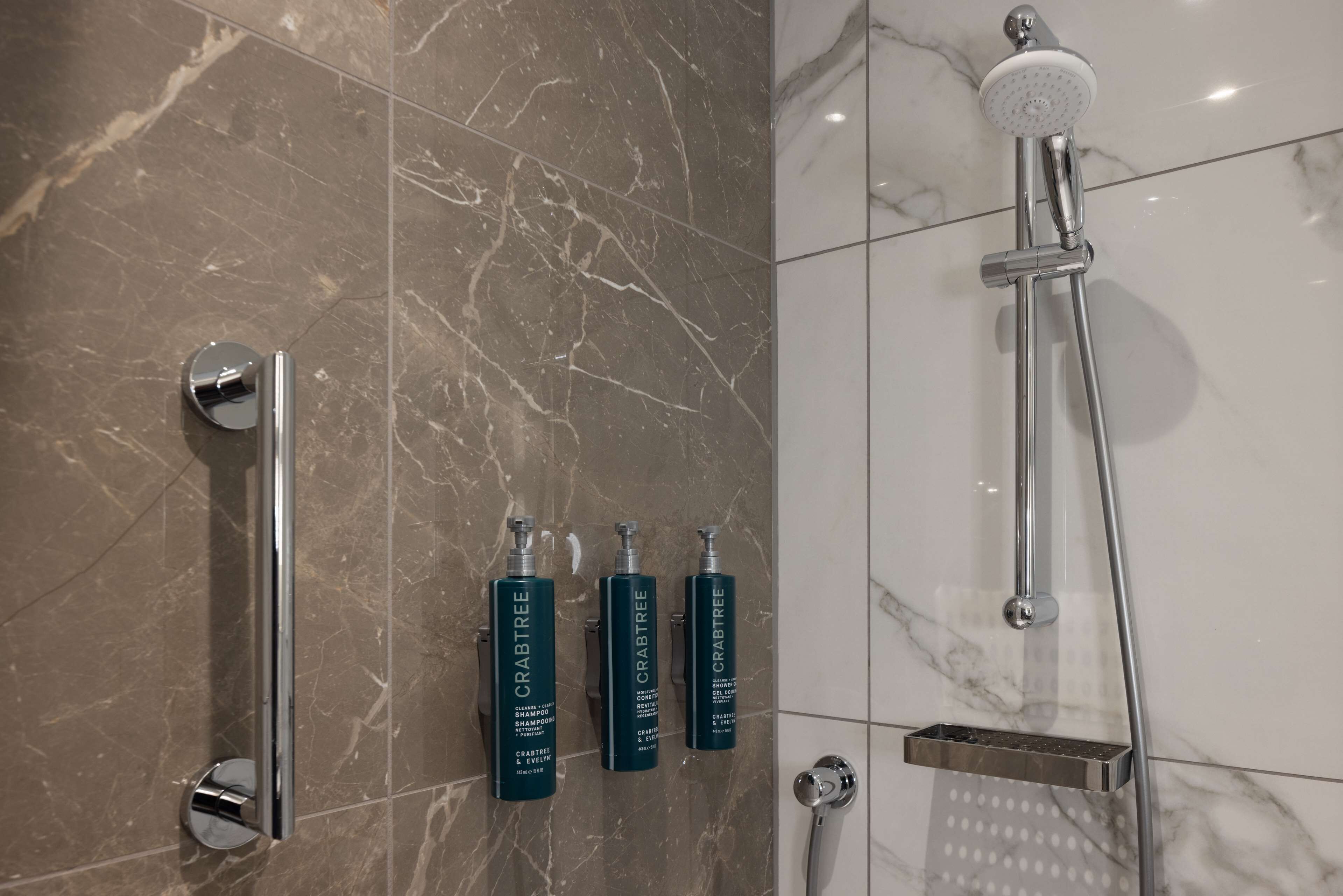 A bathroom with a marble floor, a shower with a handheld showerhead, and three bottles of Crabtree shampoo.