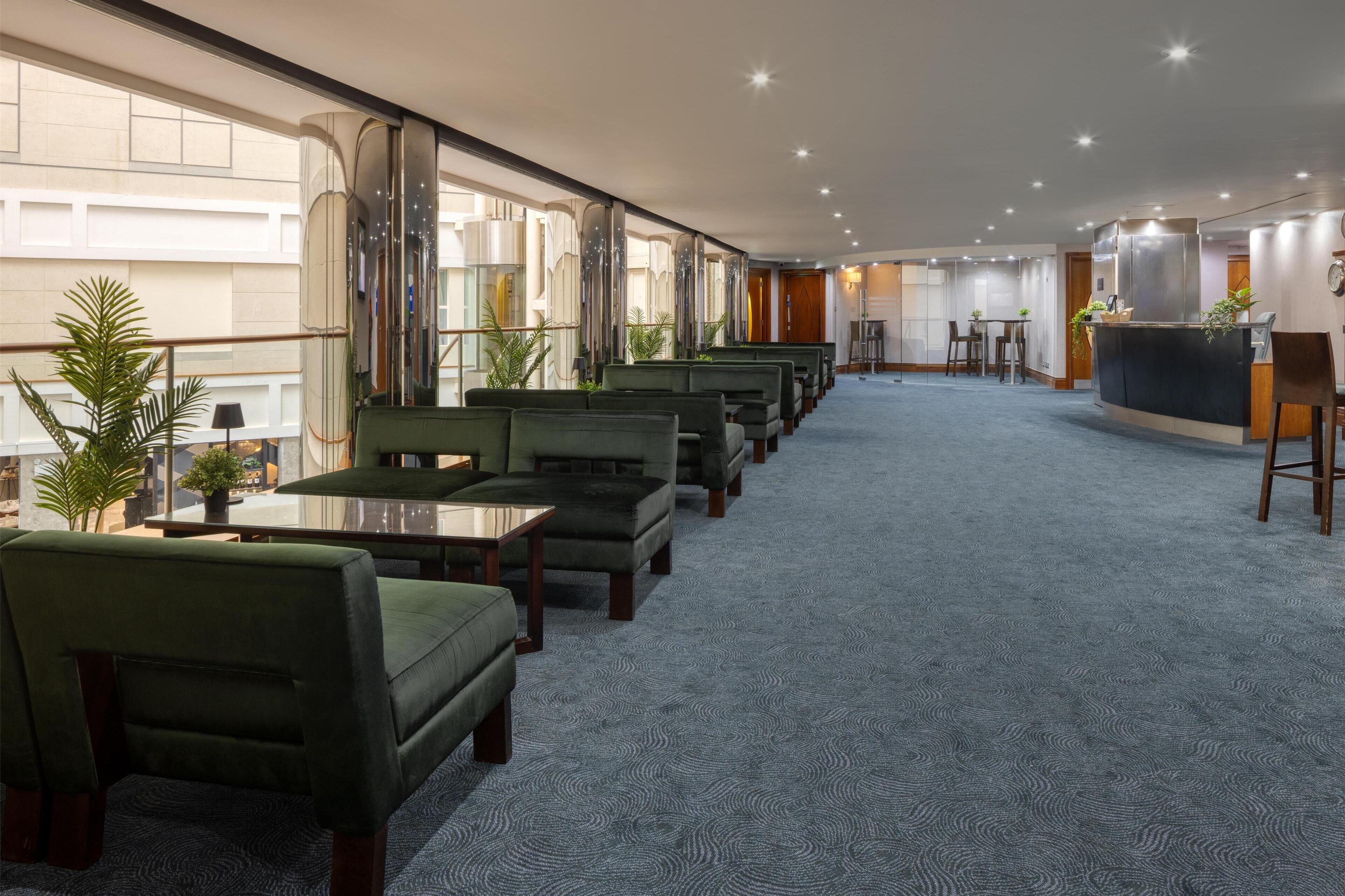 Un amplio vestíbulo de hotel con un largo mostrador de recepción, varias zonas de estar con sillones tapizados en verde y un amplio muro con ventanas que proporciona luz natural.