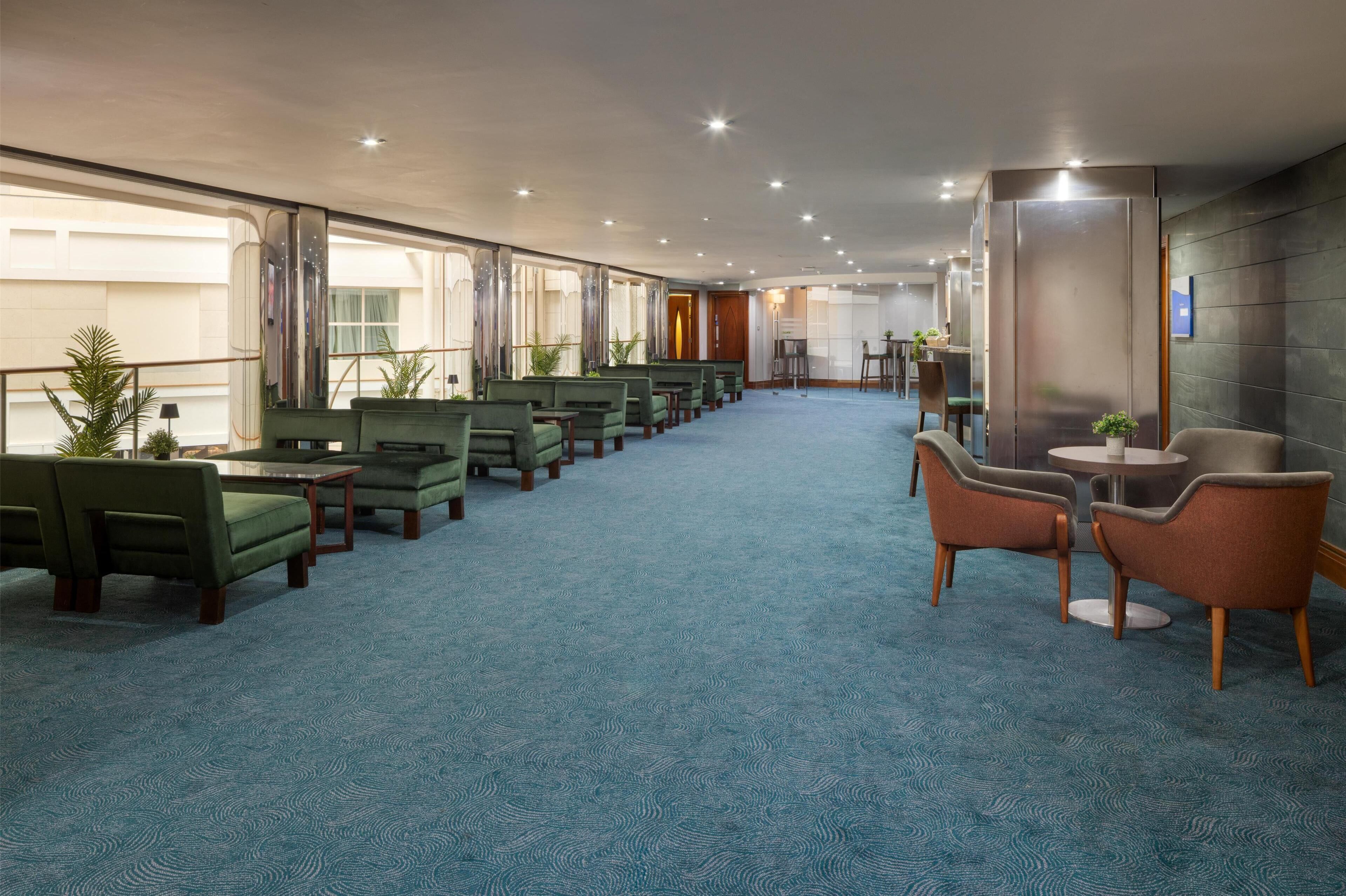 A hotel lobby with rows of green sofas and armchairs, a blue carpet, and a view of the city through large windows.
