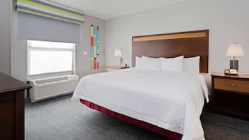 A hotel room with a large bed, a desk, a window with blinds, a wall-mounted air conditioner, and a bedside table with a lamp.