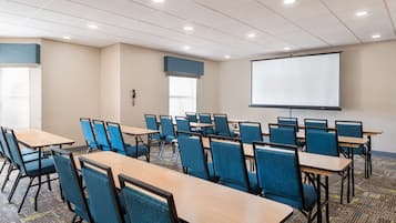 A conference room with rows of tables and chairs, a projector screen, and a phone on the wall.