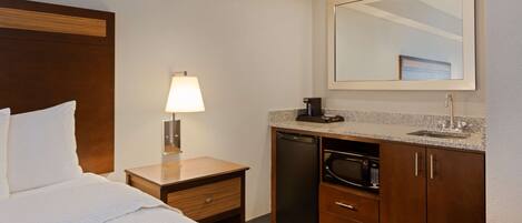 A hotel room with a bed, a nightstand with a lamp, a microwave, a sink, and a mirror.