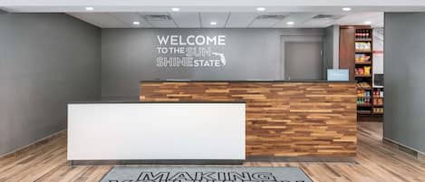 Un comptoir d’accueil avec un panneau décoratif en bois, une pancarte de bienvenue indiquant « Sunshine State », et un tapis au sol aux motifs inspirants.