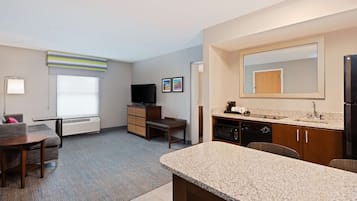 Habitación de hotel compacta con kitchenette, zona de estar con sofá y mesa, televisor de pantalla plana, espejo grande y ventana con persianas.