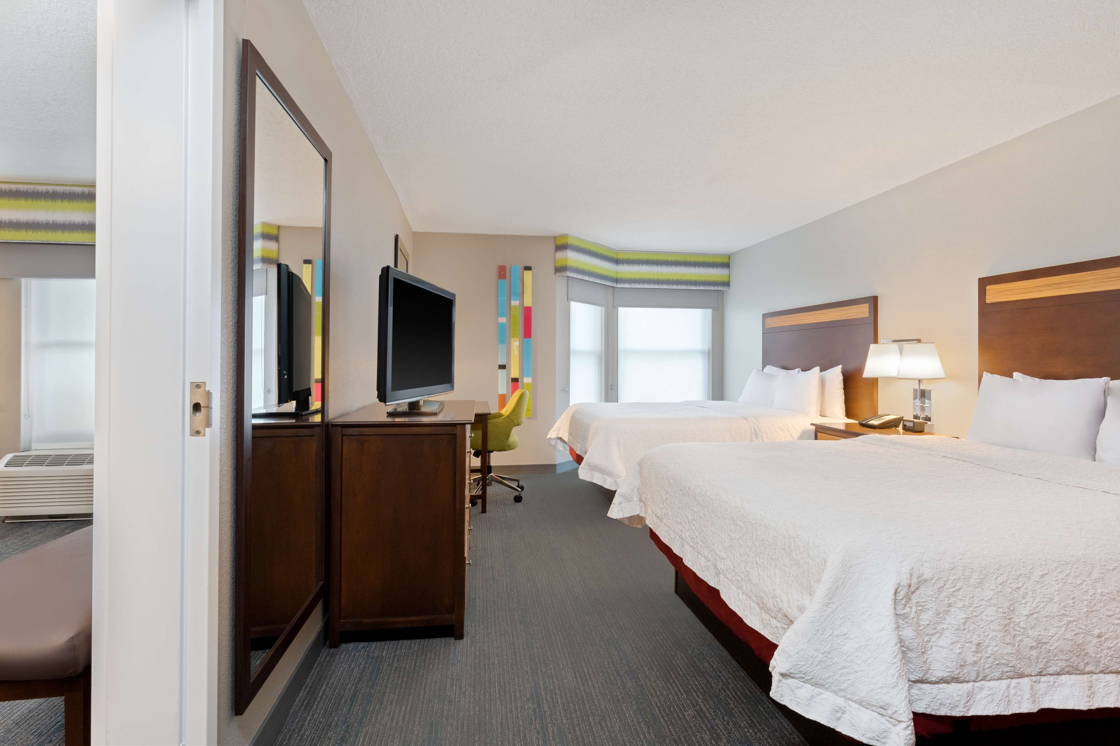 A hotel room with two beds, a desk with a TV, a chair, a window, and a lamp.