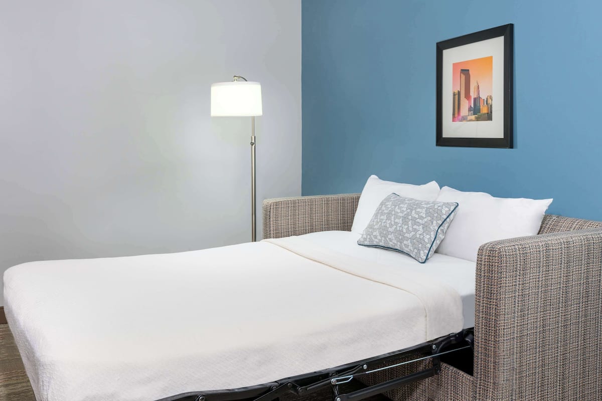 Un lit simple avec un drap blanc, un oreiller gris et un tableau représentant un paysage urbain accroché au mur.
