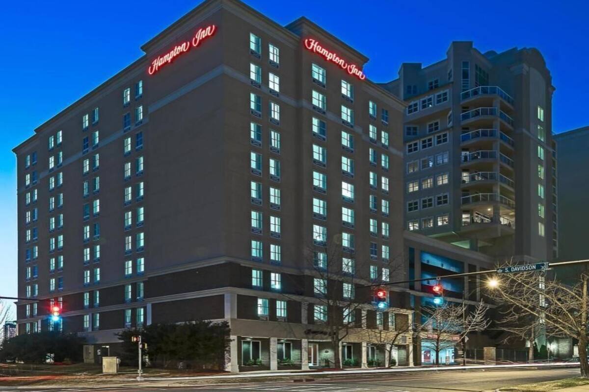 Un hôtel à plusieurs étages, reconnaissable à son grand panneau « Hampton Inn », doté de nombreux balcons et donnant sur une rue avec des feux de signalisation.