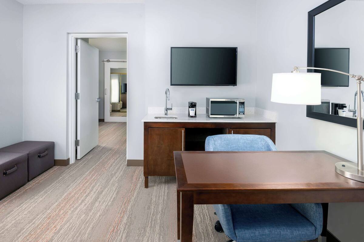 Une chambre d’hôtel avec un bureau en bois, un téléviseur à écran plat fixé au-dessus, un four à micro-ondes, une machine à café, une table de chevet et une chaise.