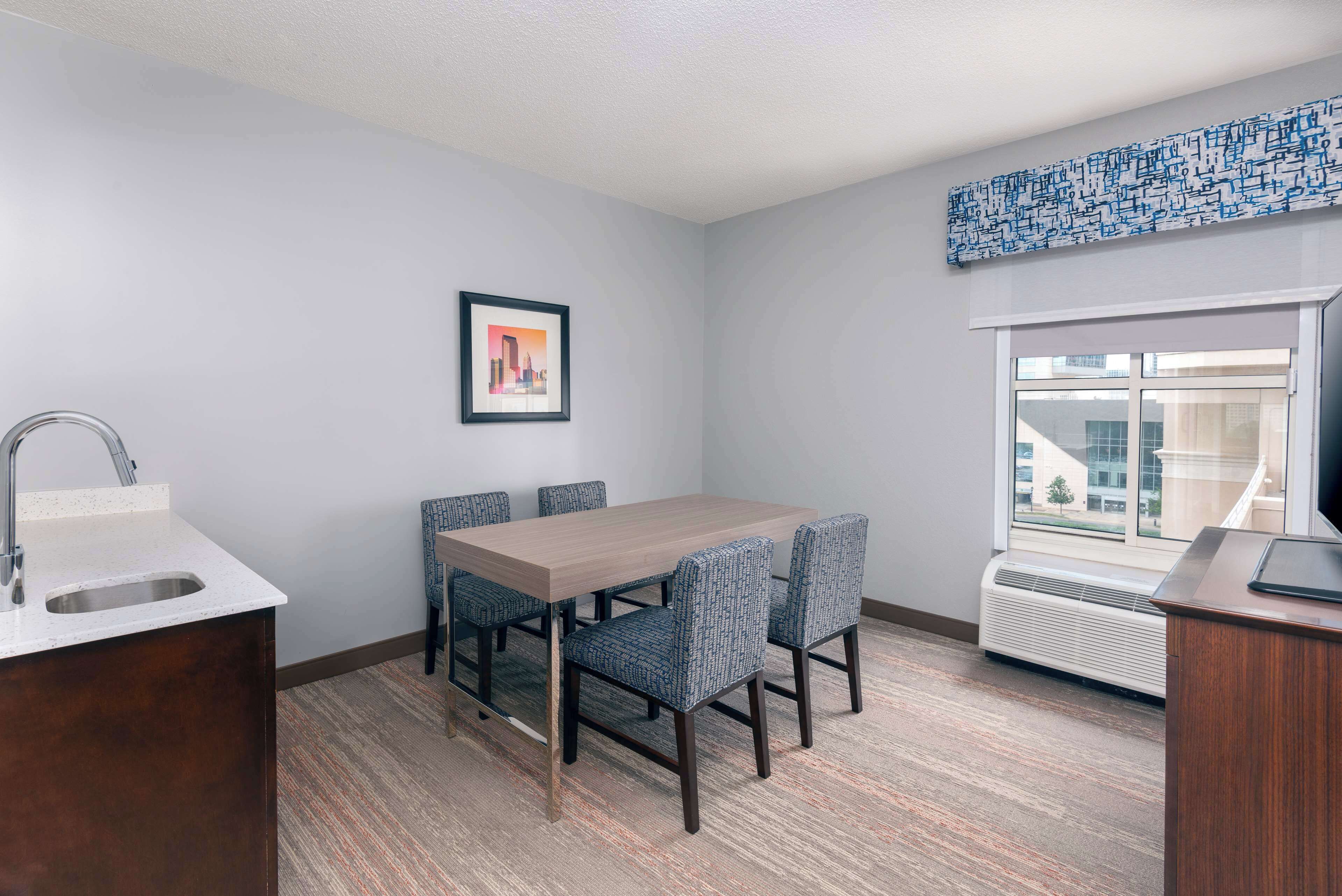 Una habitación de hotel pequeña y moderna con una mesa de comedor de madera, cuatro sillas, un lavamanos empotrado y un ventanal con vistas a la ciudad.