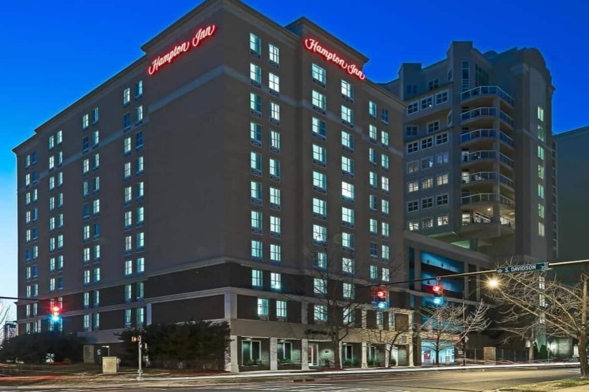 Un hôtel à plusieurs étages, reconnaissable à son grand panneau « Hampton Inn », doté de nombreux balcons et donnant sur une rue avec des feux de signalisation.