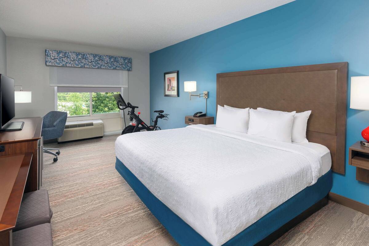 Une chambre d’hôtel avec un grand lit, un bureau avec une chaise, un vélo et une fenêtre donnant sur des arbres.