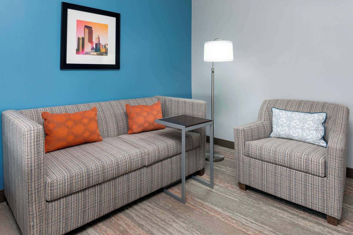 Une pièce avec un mur bleu, un canapé gris à motifs agrémenté de coussins orange, un fauteuil gris à motifs avec un coussin bleu, une petite table et un lampadaire.