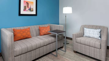 Une pièce avec un mur bleu, un canapé gris à motifs agrémenté de coussins orange, un fauteuil gris à motifs avec un coussin bleu, une petite table et un lampadaire.