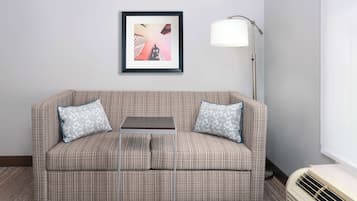 A room with a patterned sofa, two pillows, a small table, a floor lamp, and a wall-mounted picture.