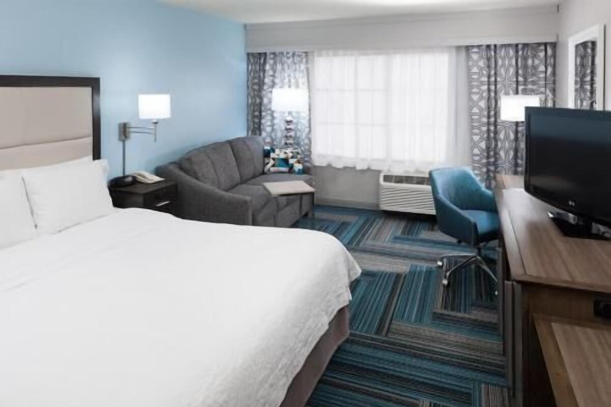 A hotel room with a bed, a desk with a TV, a chair, a desk lamp, and a window with curtains.