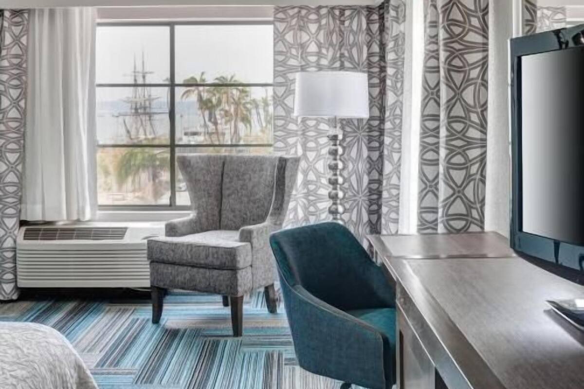 A hotel room with a large window, a chair, a desk, and a television.