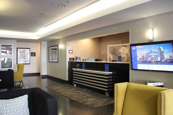 Ein Hotel-Empfangsbereich mit einer Rezeption, einem Fernseher, der das Hampton Inn Logo zeigt, gerahmten Kunstwerken an den Wänden und einem gelben Sessel.