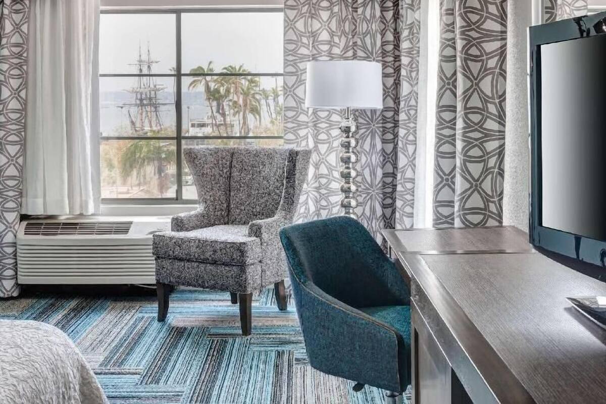 A hotel room with a large window, a patterned wall, a chair, a desk, and a TV.