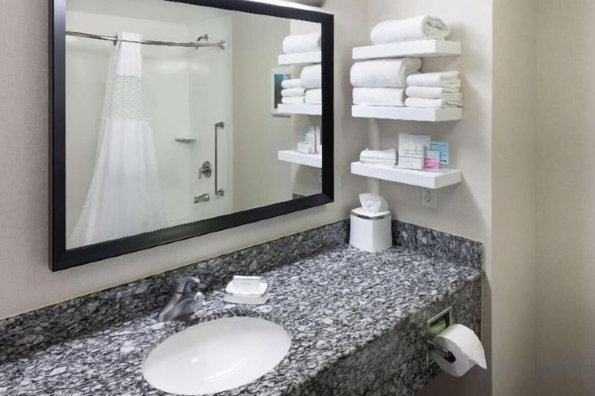A bathroom with a granite countertop, a white sink, a mirror, and shelves with towels and toiletries.