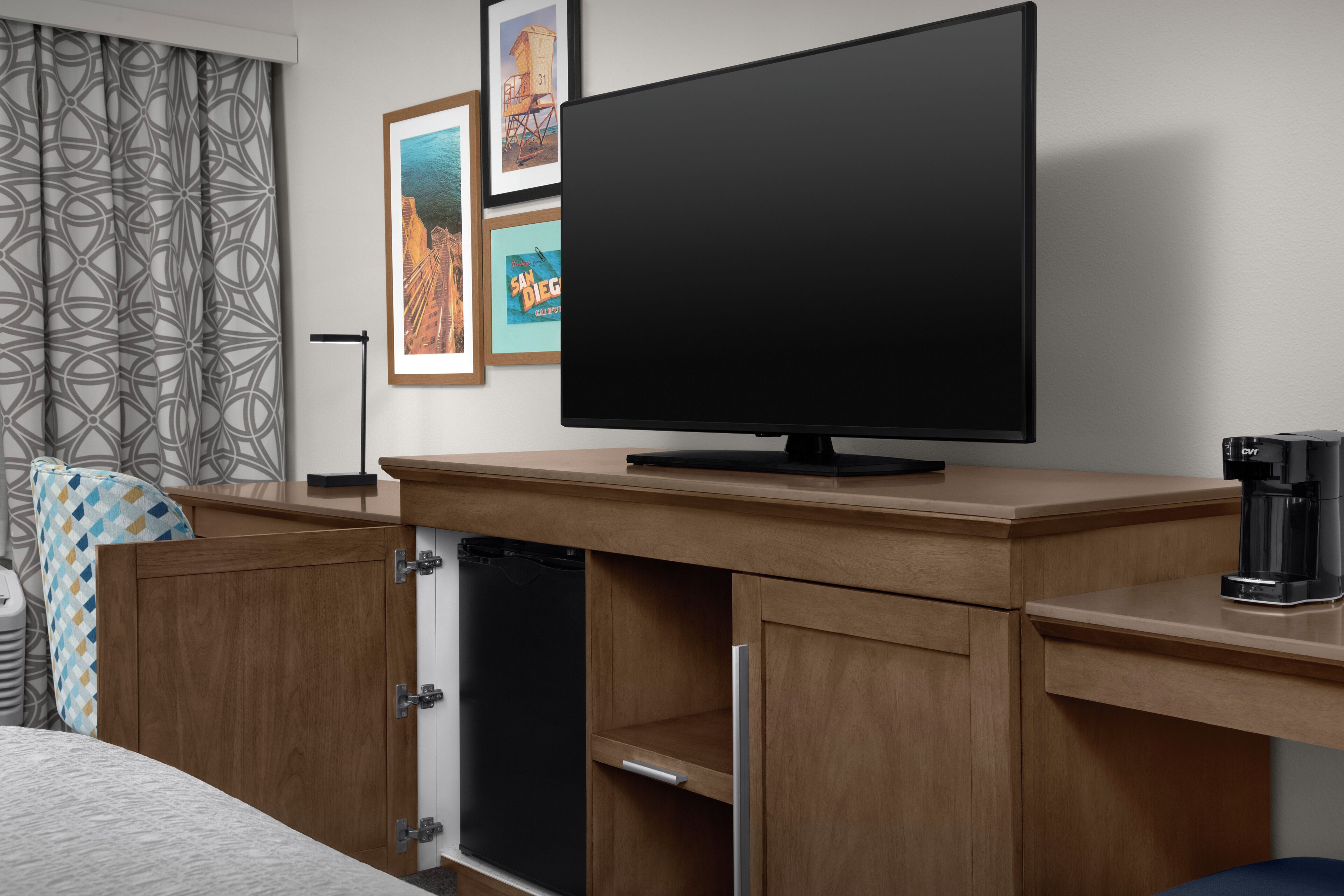Habitación de hotel con televisión de pantalla plana sobre un mueble de madera, cafetera y refrigerador.