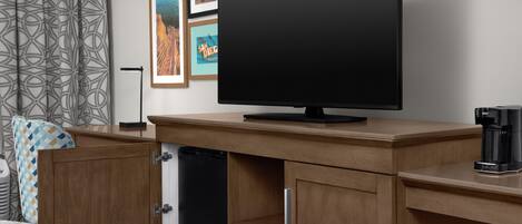 Habitación de hotel con televisión de pantalla plana sobre un mueble de madera, cafetera y refrigerador.
