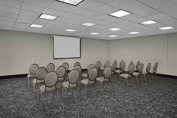 Un salón de conferencias con pantalla de proyector, filas de sillas y suelo alfombrado.