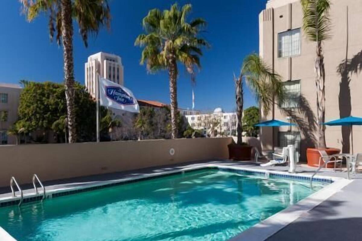 A swimming pool with lounge chairs and umbrellas, surrounded by palm trees and buildings.
