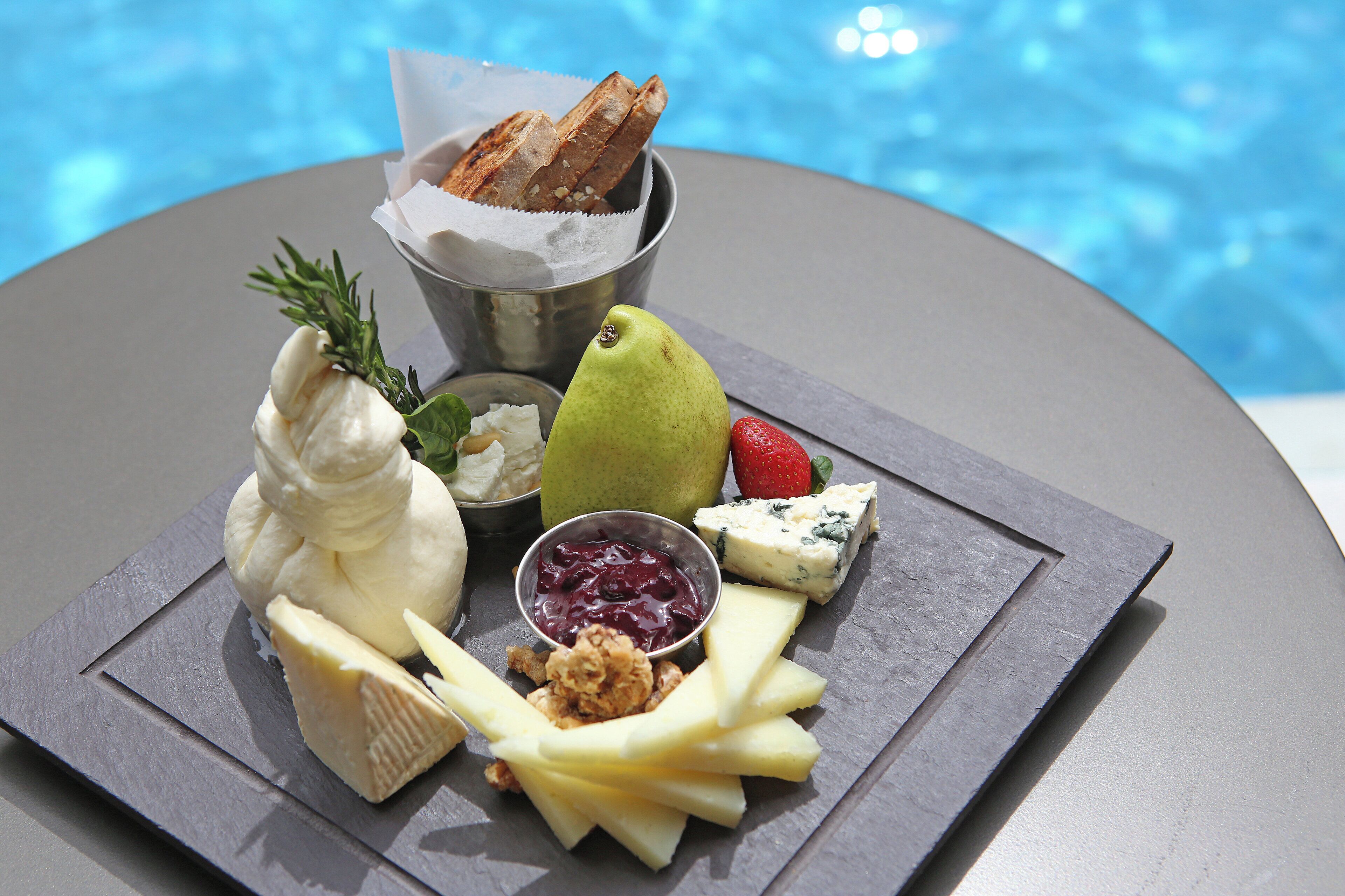 A cheese board with various cheeses, fruits, and nuts by a poolside.