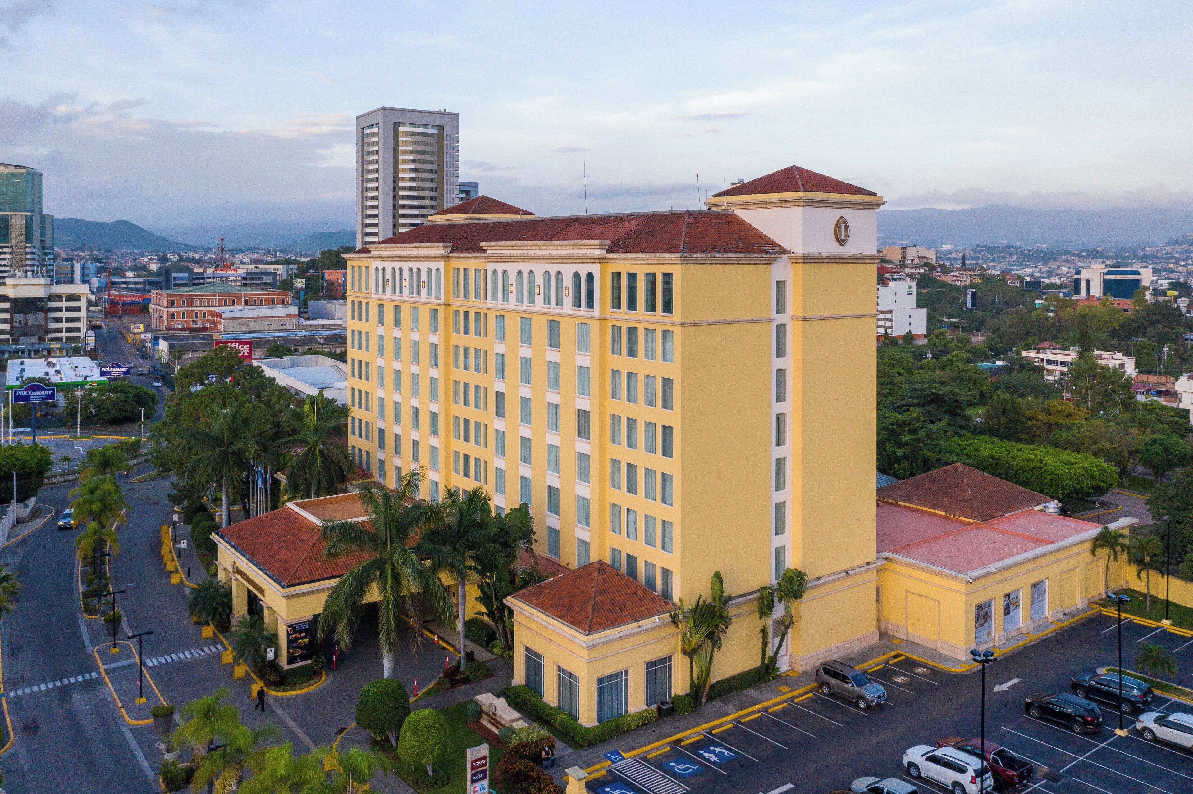 A multi-story hotel with a yellow facade and red-tiled roofs, surrounded by palm trees and a parking lot.