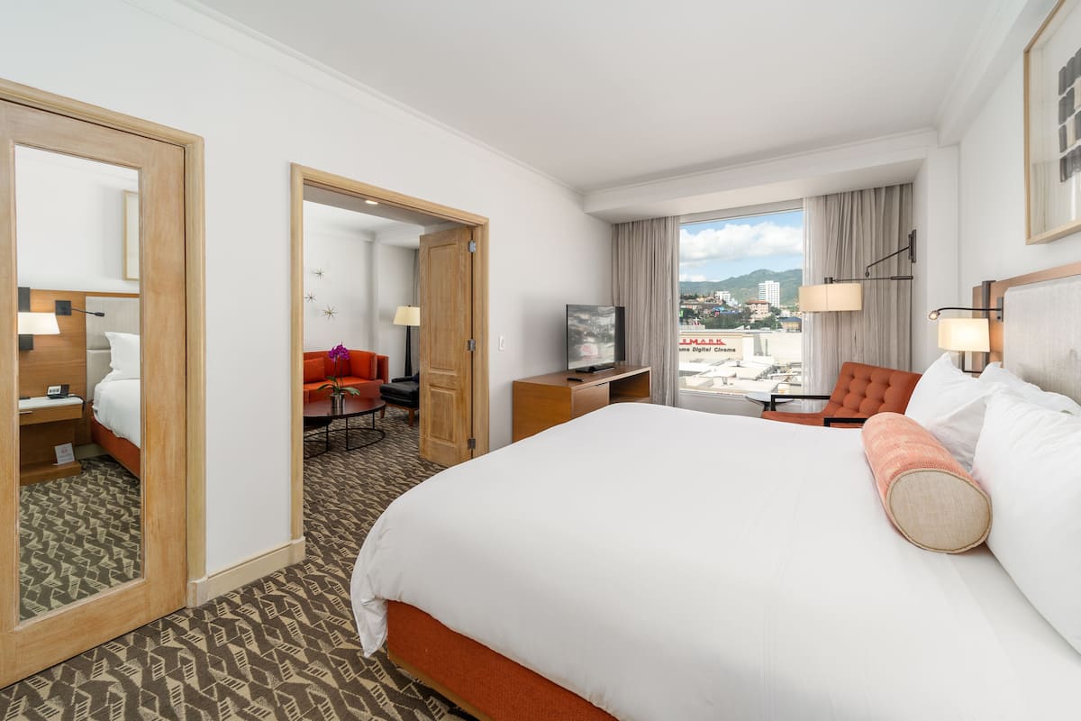 Ett hotellrum med en stor säng, ett skrivbord med en tv, en stol och utsikt över staden.