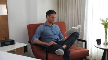 A man sitting on a red chair in a hotel room, reading a book. There is a small table with a plant and a black mug on it.