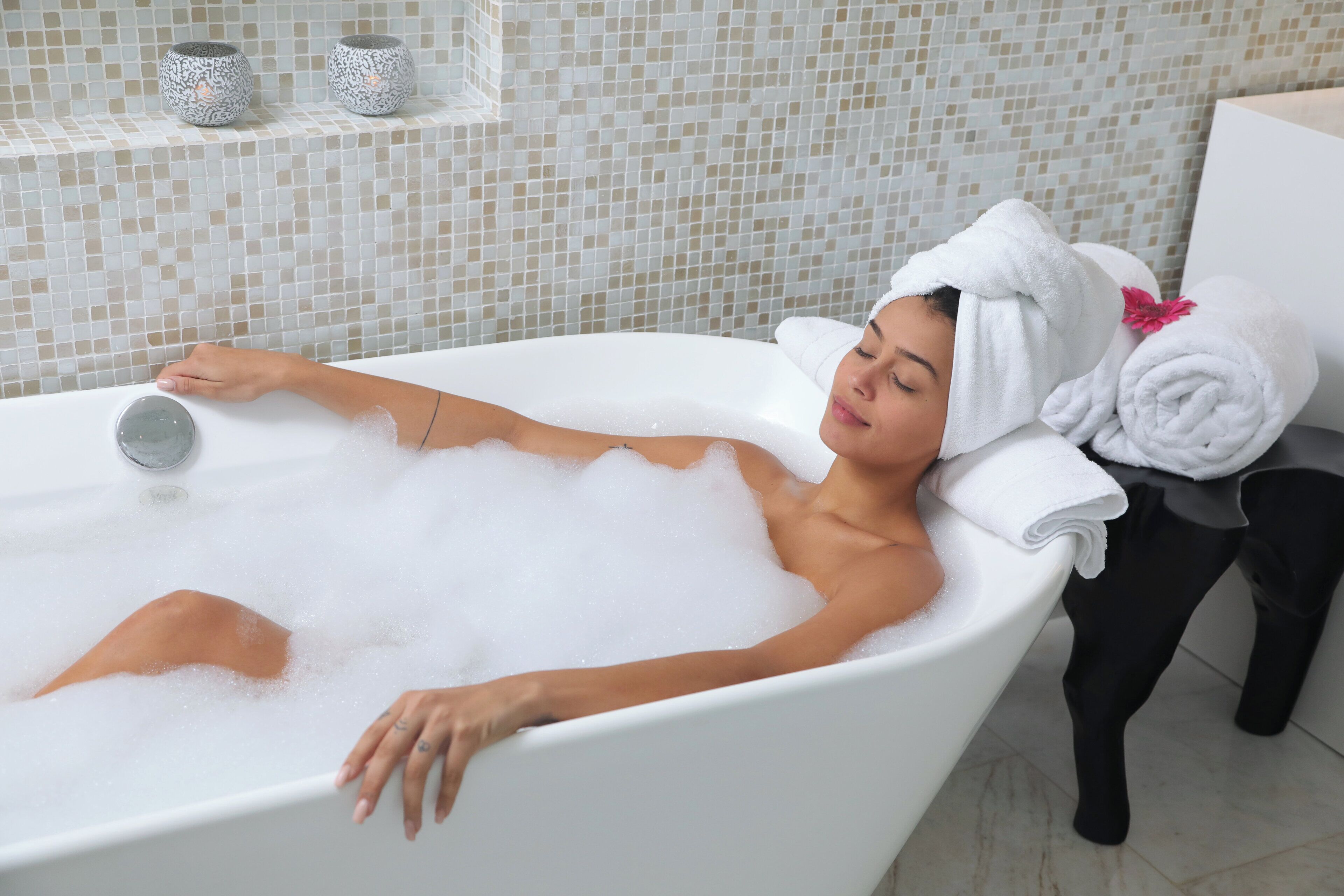 Una persona relajándose en una bañera llena de burbujas, con una toalla en la cabeza y otra en el suelo cerca de la bañera.