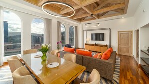 Una habitación de hotel moderna con techo de madera, ventanas amplias, zona de comedor con mesa de vidrio y sala de estar con sofá y televisor.