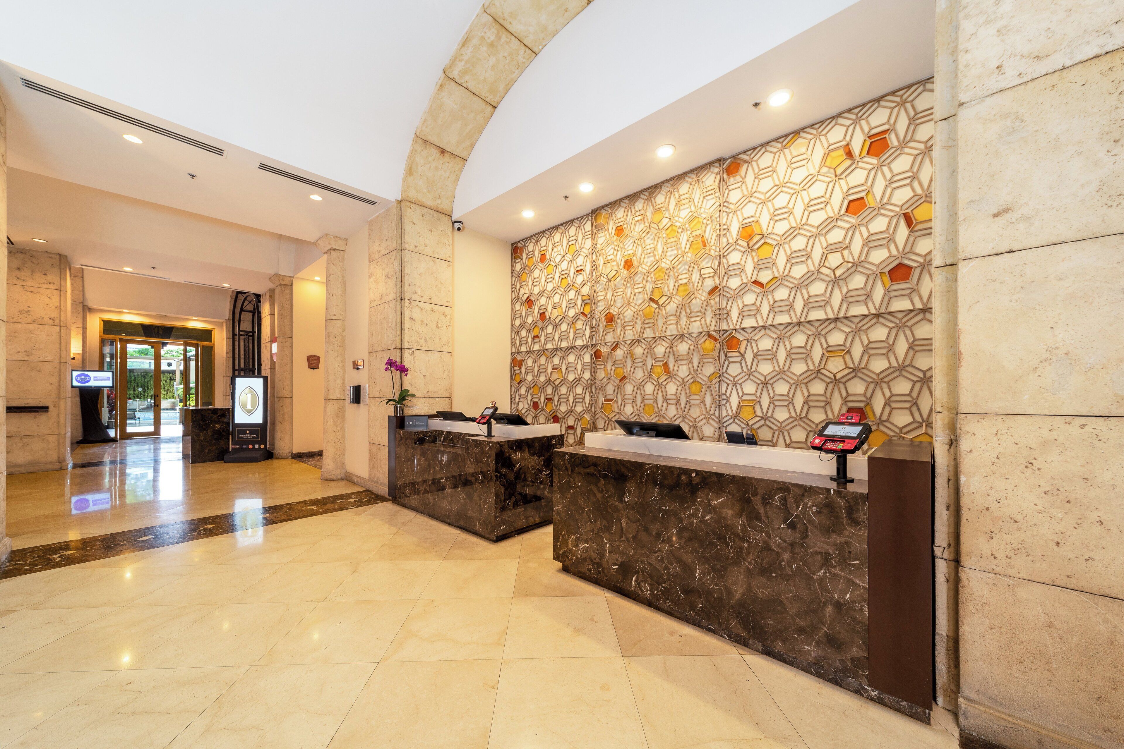 Zona de recepción de hotel con mostradores de mármol, pared con patrón geométrico y vista despejada a la entrada.