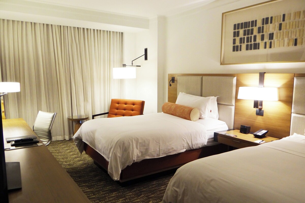 Habitación de hotel con una cama grande, una cama más pequeña, un escritorio con lámpara, una silla y un televisor instalado en la pared.