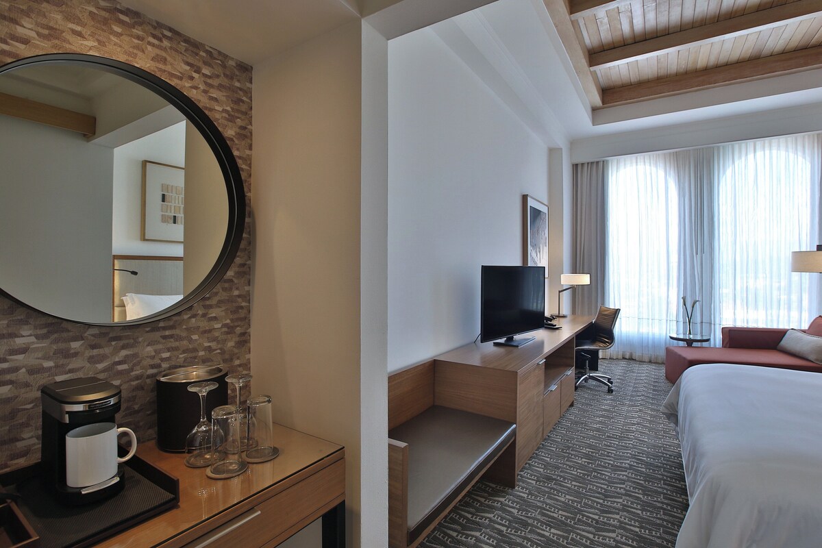 Habitación de hotel con escritorio de madera, televisor, cama y un espejo grande.