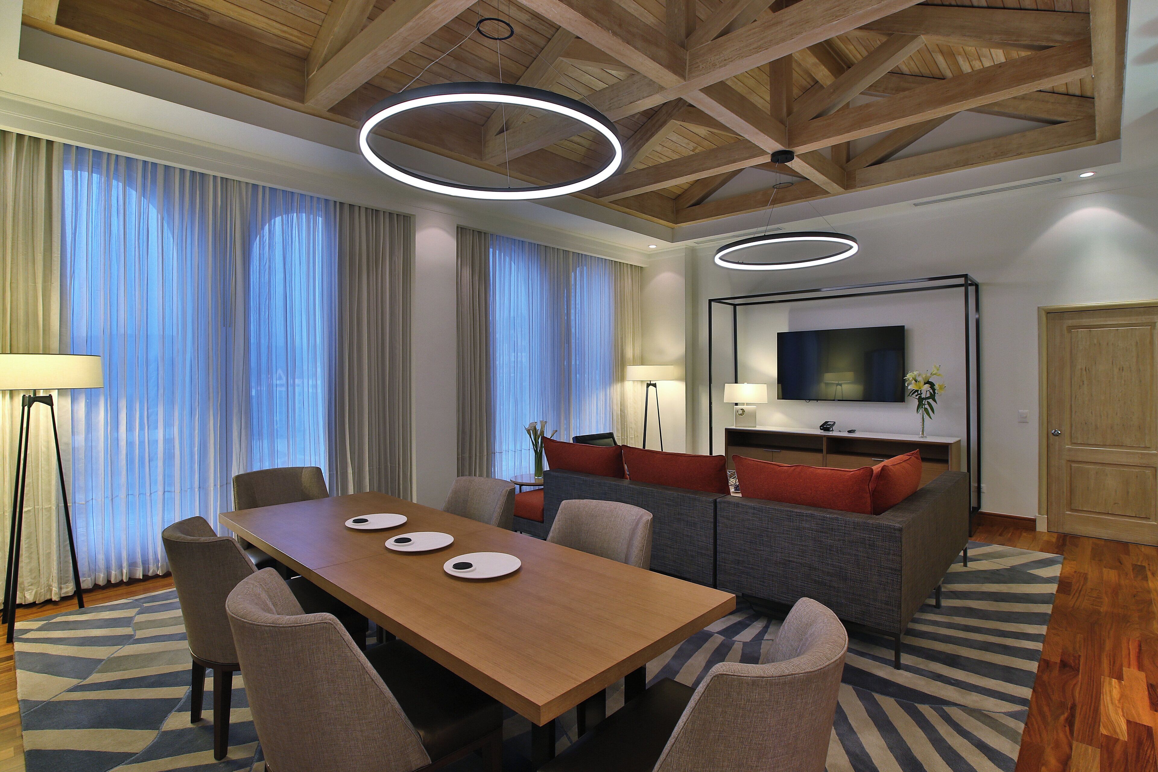 A modern hotel room with a wooden dining table, comfortable seating, a flat-screen TV, and a large window with curtains.