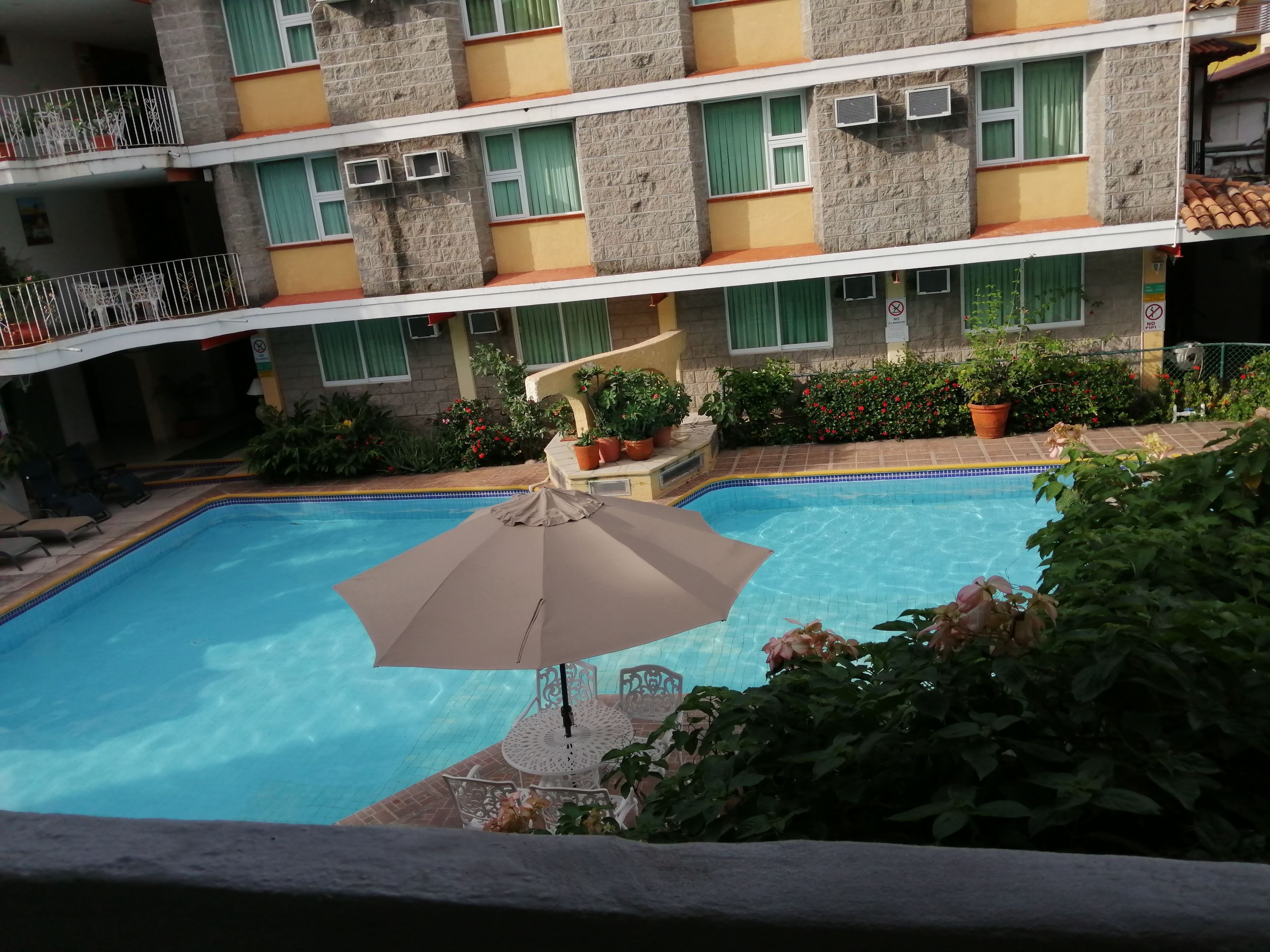 A swimming pool with a lounge area, surrounded by potted plants and a building with balconies.