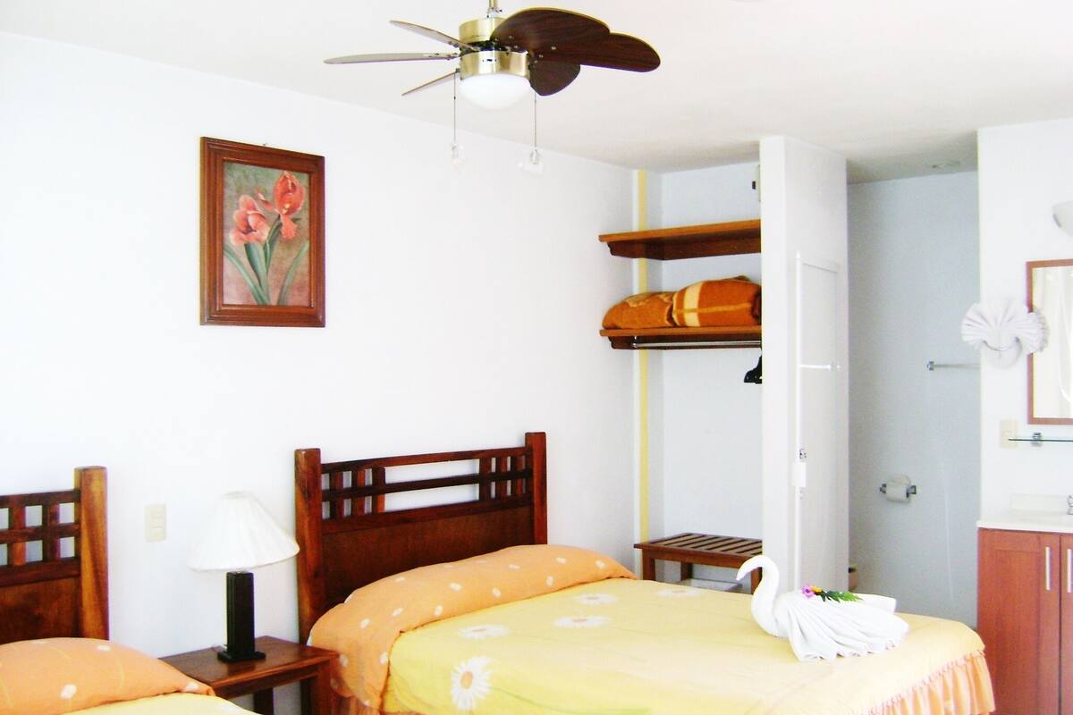 Hotelkamer met twee houten bedden, een plafondventilator, een schilderij, een plank met een koffer, en een badkamer met spiegel en handdoekhouder.