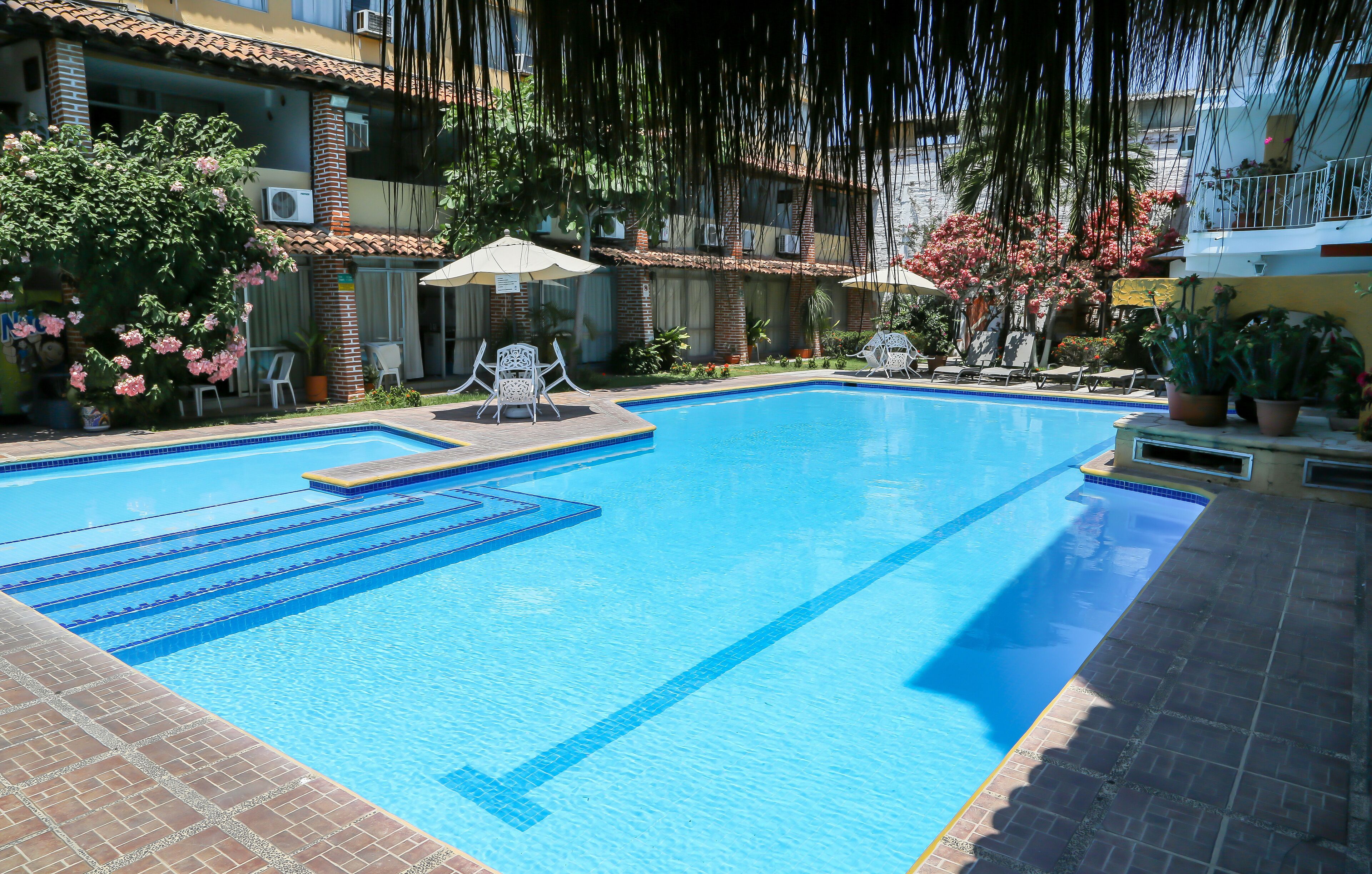 A swimming pool with clear blue water, surrounded by lounge chairs and umbrellas, with a brick building and greenery in the background.