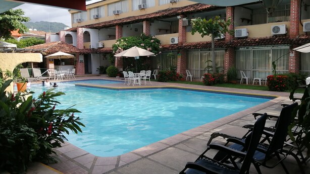 A swimming pool with lounge chairs and umbrellas, surrounded by a building with multiple air conditioning units and a view of mountains in the background. - Hotel Vallartasol (Puerto Vallarta)