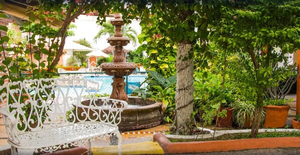Une fontaine, un banc en fer forgé blanc et des plantes en pot dans un espace aménagé en jardin.