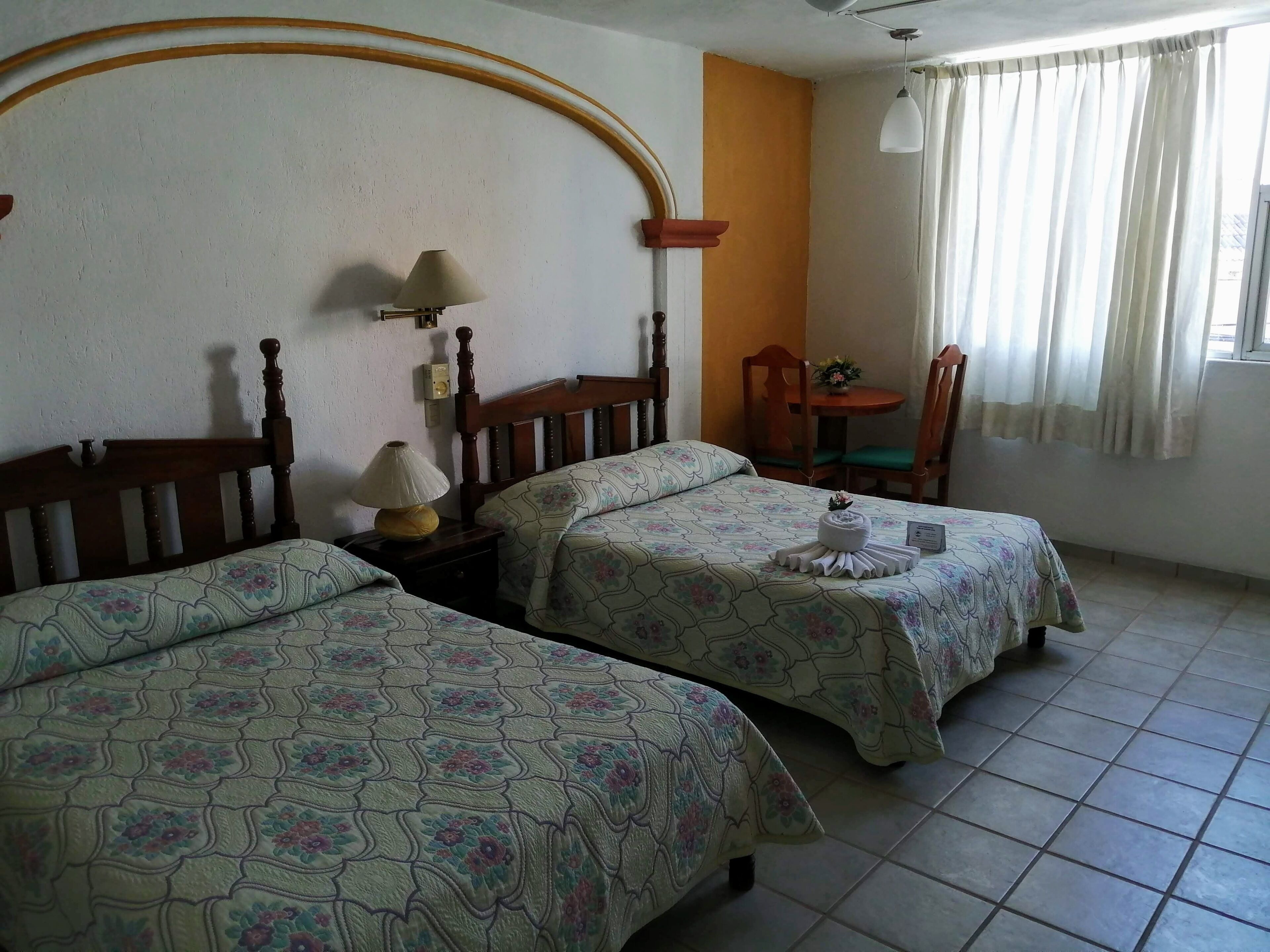 Una habitación con dos camas, una mesita con una lámpara, una silla y una ventana con cortinas.