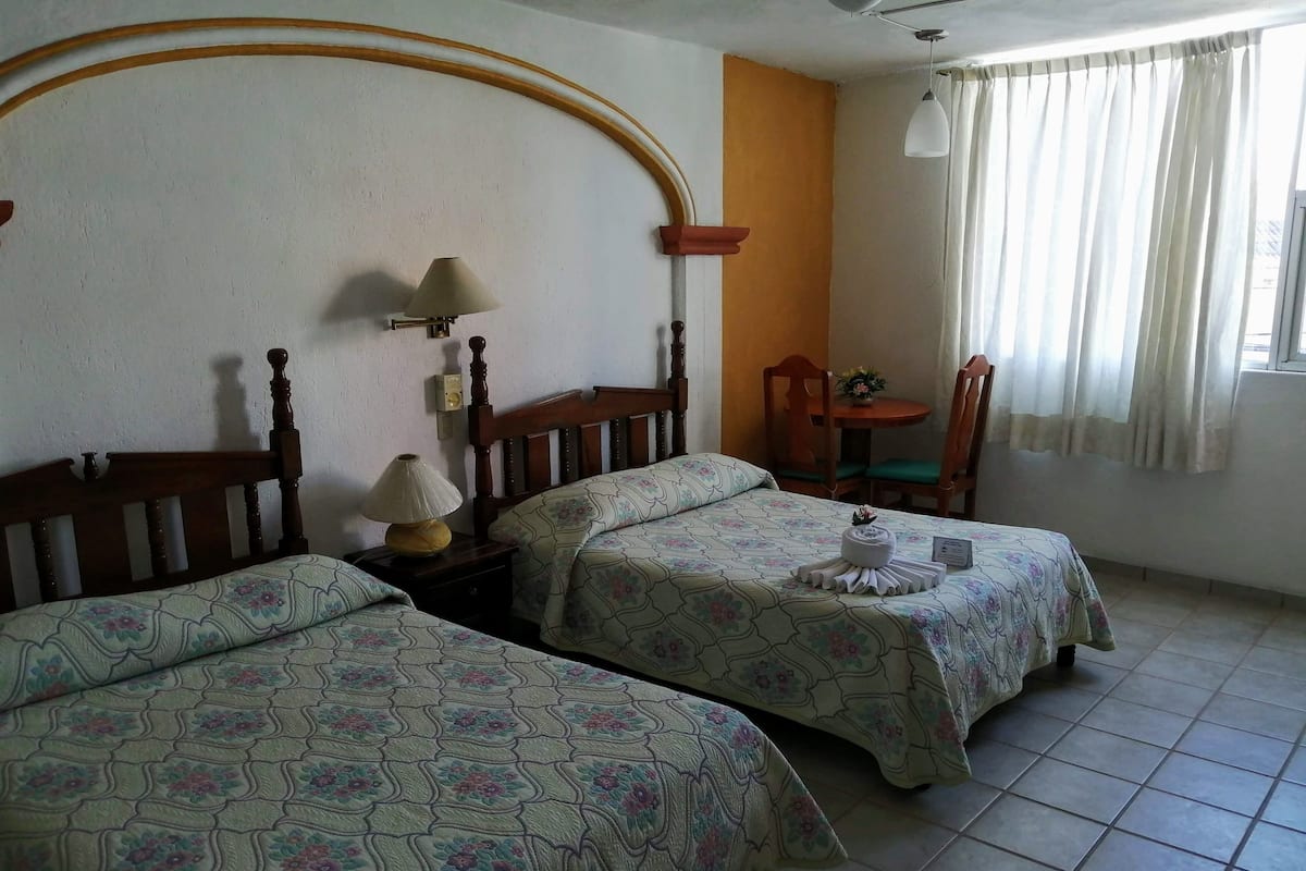 Ein Zimmer mit zwei Betten, einem kleinen Tisch mit Lampe, einem Stuhl und einem Fenster mit Vorhängen.