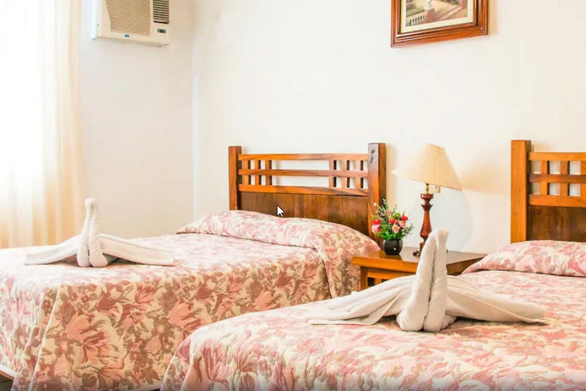 A hotel room with two beds, each with a floral bedspread and wooden headboards. There is a nightstand with a lamp and a vase of flowers between the beds.