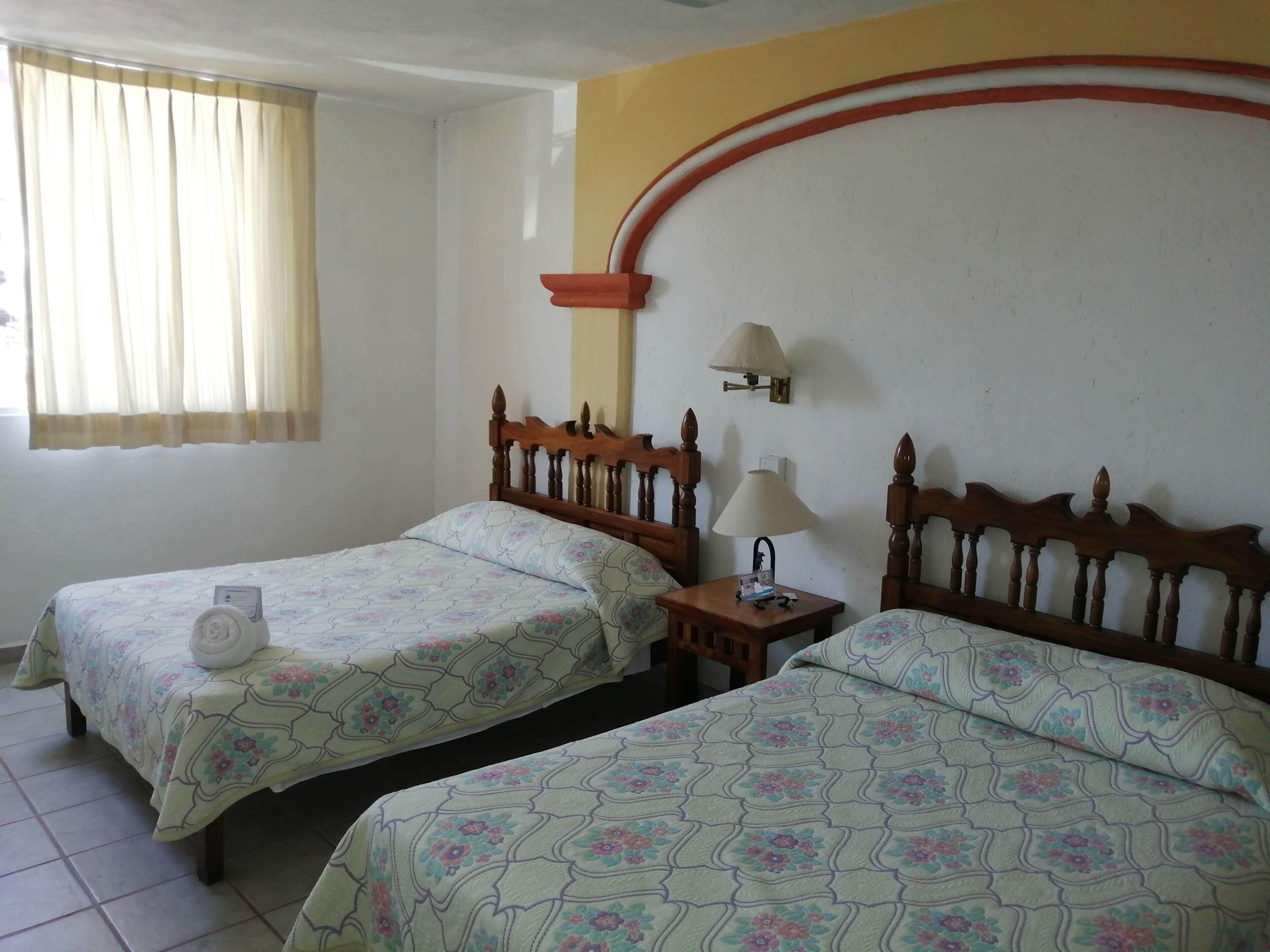 A room with two single beds, a nightstand with a lamp, a wall-mounted shelf, and a window with curtains.