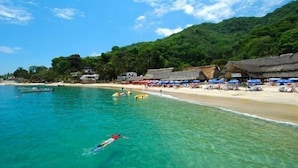 A beach with clear turquoise water, a swimmer, and thatched-roof huts along the shore. - Hotel Vallartasol (Puerto Vallarta)
