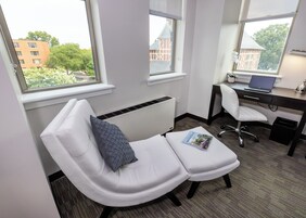 A room with a massage chair, a desk with a laptop, and large windows offering a view of buildings and trees.