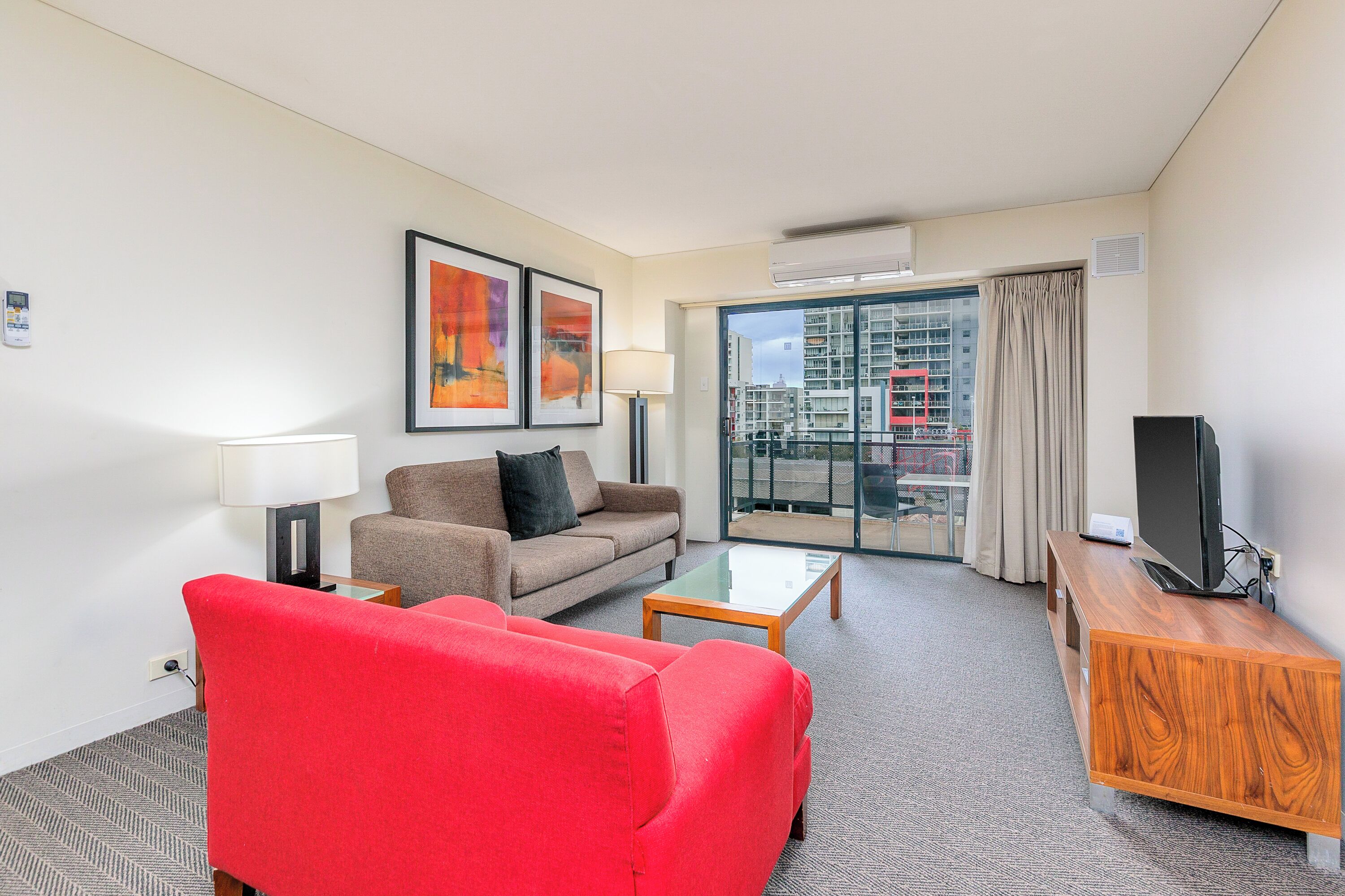 Ein Hotelzimmer mit einem roten Sofa, einem braunen Sofa, einem Glastisch, einer Holz-TV-Konsole und einem großen Fenster mit Blick auf Gebäude.