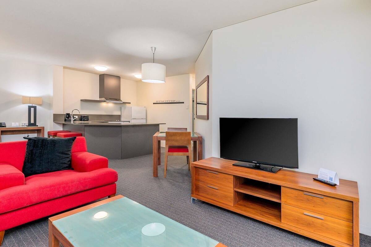 A modern hotel room with a red sofa, a glass coffee table, a wooden TV stand, a flat-screen TV, a kitchen area with a stove and microwave, and a small dining table.